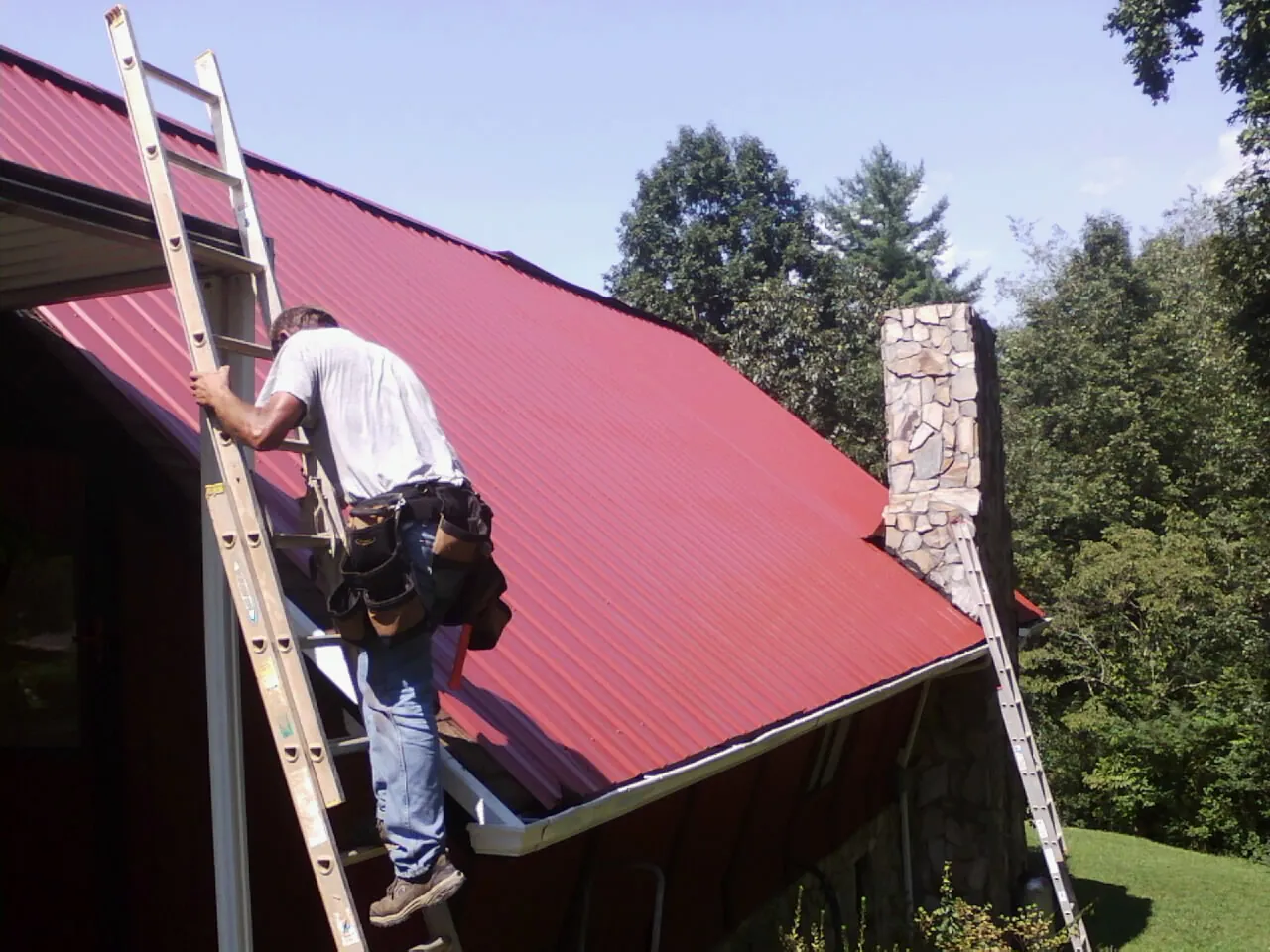 Roofer on a ladder beside a red metal roof and stone chimney — Soffit & Fascia Repair by Roofing Brothers in Shepherd